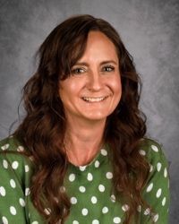 Smiling woman with long brown hair wearing a green polka-dot blouse against a gray backdrop