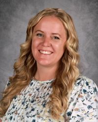 Smiling woman with long blonde hair in a patterned blouse against a gray backdrop