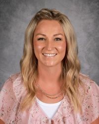 Smiling person with blonde hair wearing a pink blouse against a gray studio backdrop