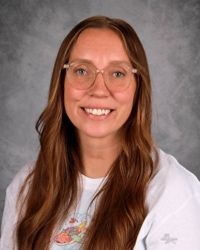 Smiling woman with glasses and long brown hair in a white shirt against a gray background