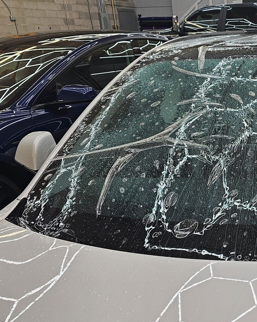 Car windshield shattered with spiderweb cracks, parked beside a dark blue car in a garage-like setting