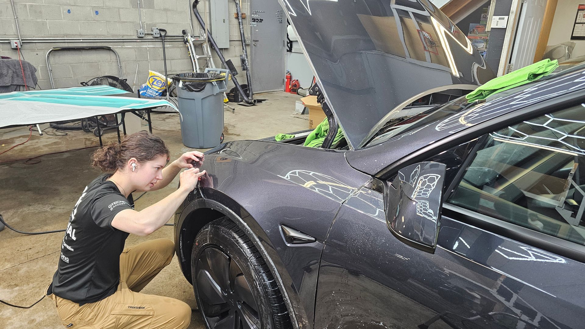 Technician wetsands the front fender of a black sports car in a garage.