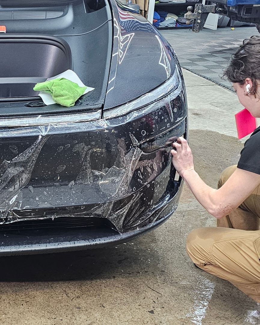 Person cleaning mud off the front bumper of a black car with a green cloth.