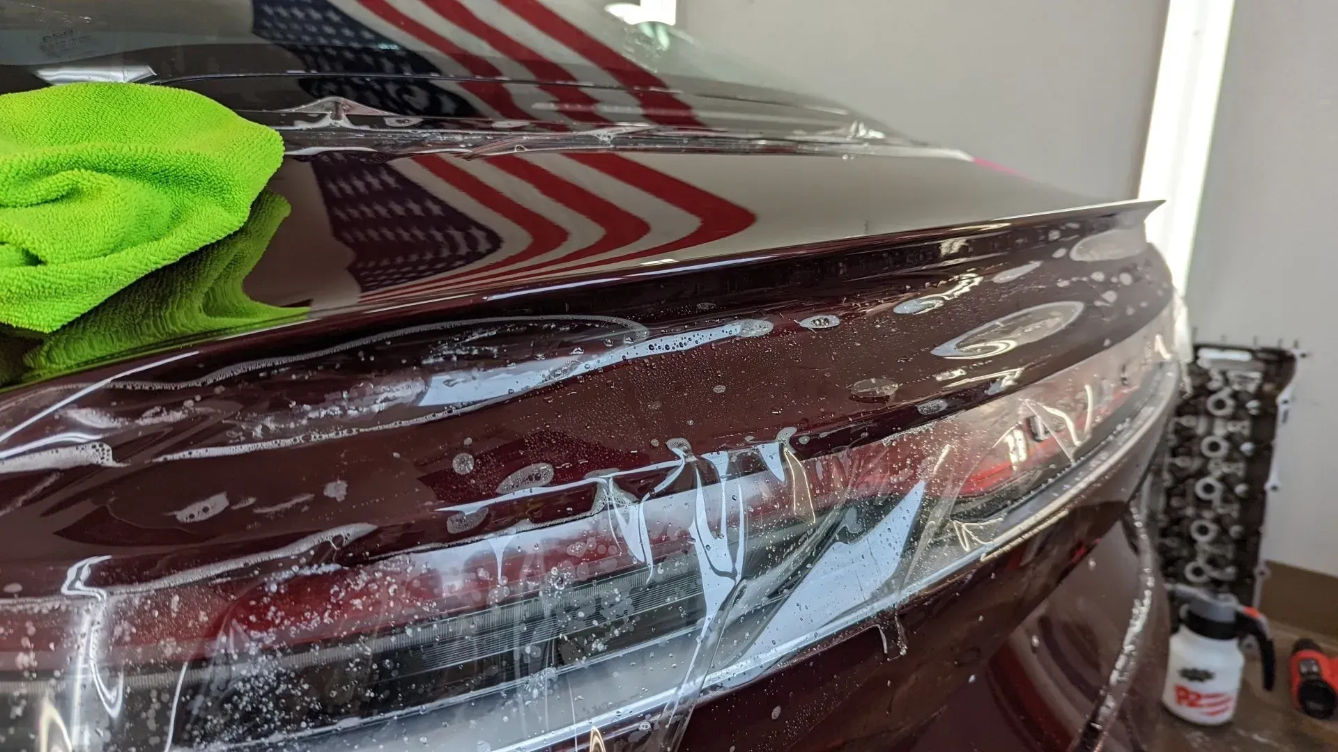 A maroon car hood wrapped in clear plastic, with a green microfiber cloth on top and an American flag reflection.