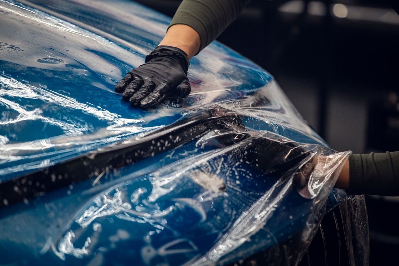 Gloved hands smoothing clear plastic wrap over a blue car hood during detailing