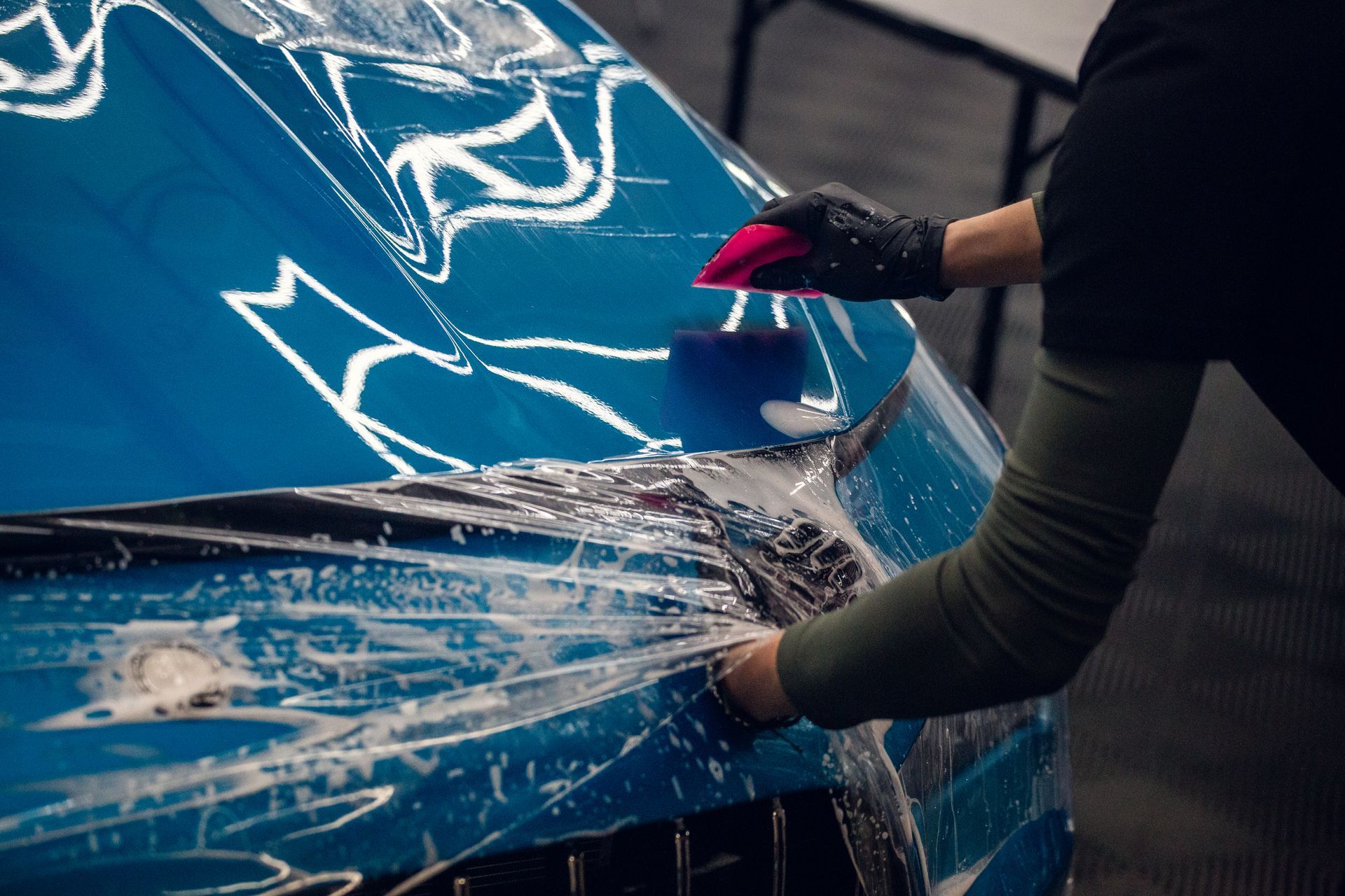 Person washing a blue car hood with water spray and a cloth