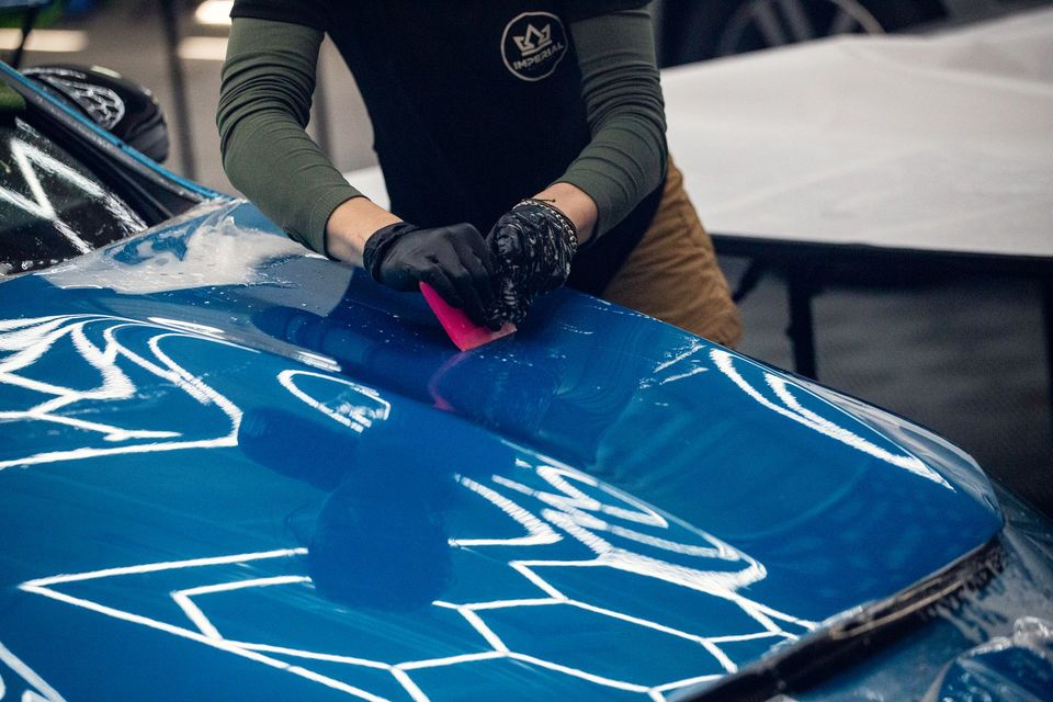 Person in gloves polishes a blue car hood with a pink cloth, showing white reflections