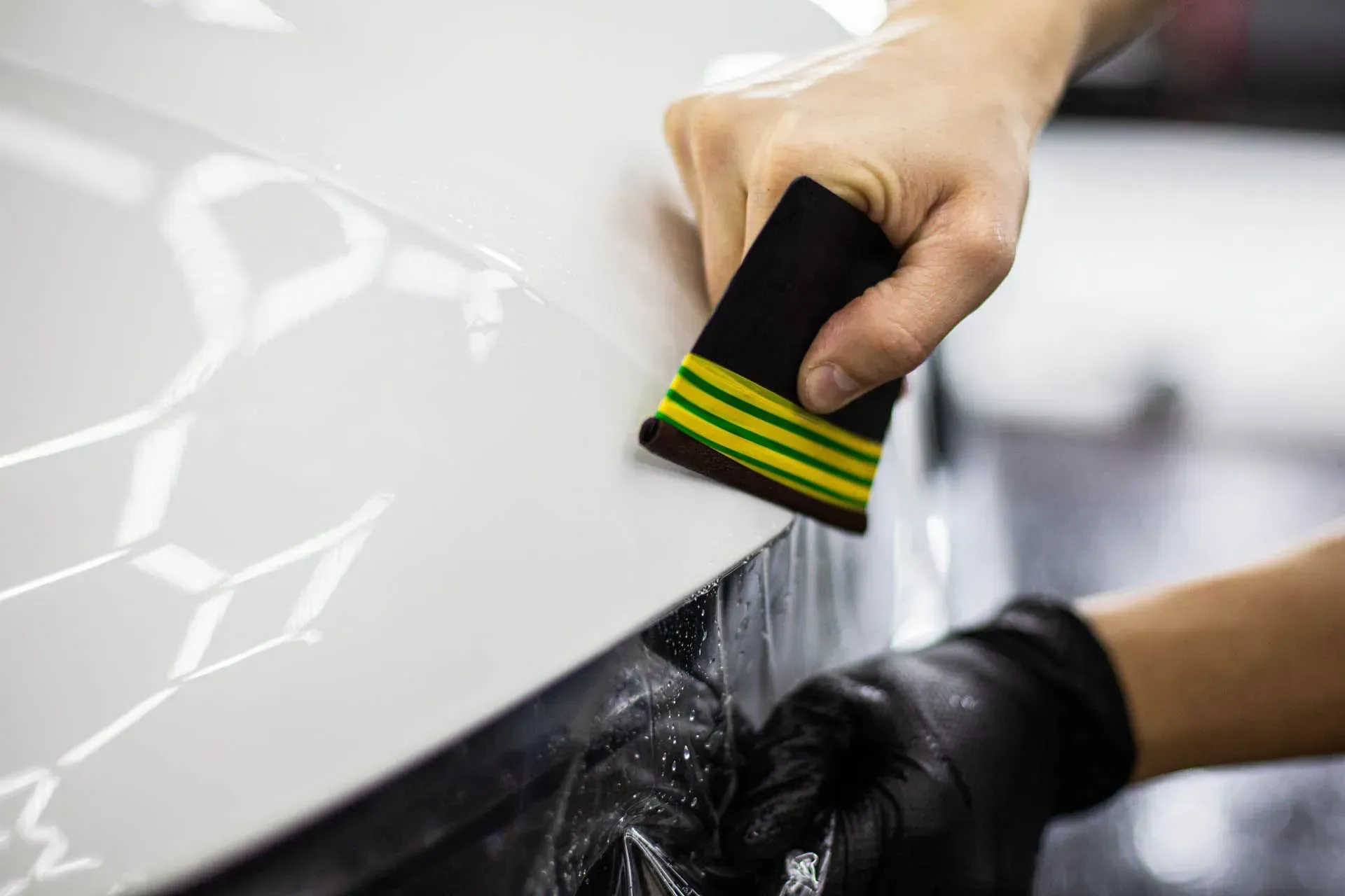 Hands using a black-and-yellow brush to scrub a white car surface with soapy water