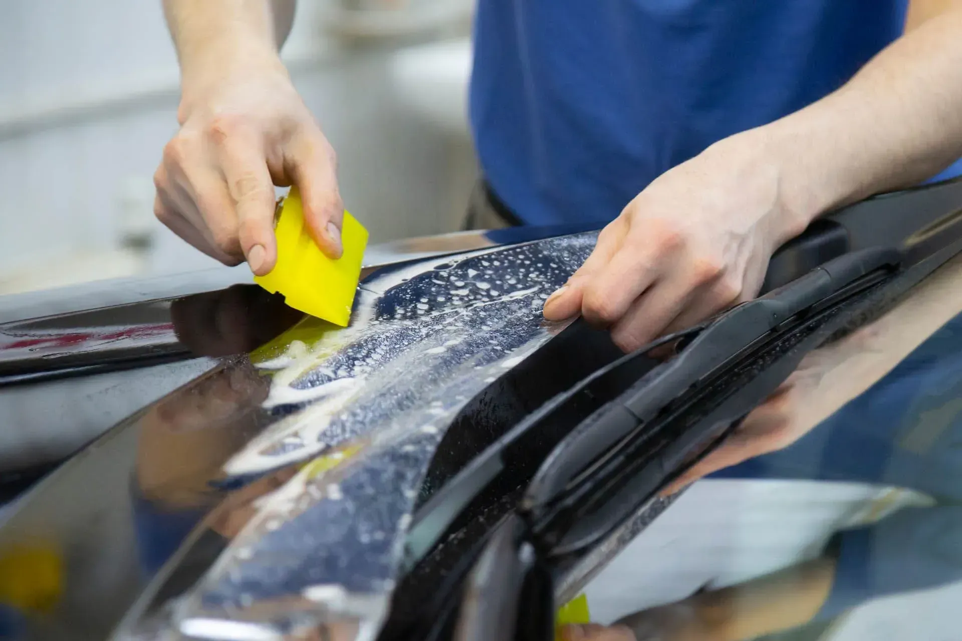 Hands scrubbing a car windshield with a yellow sponge and soapy water
