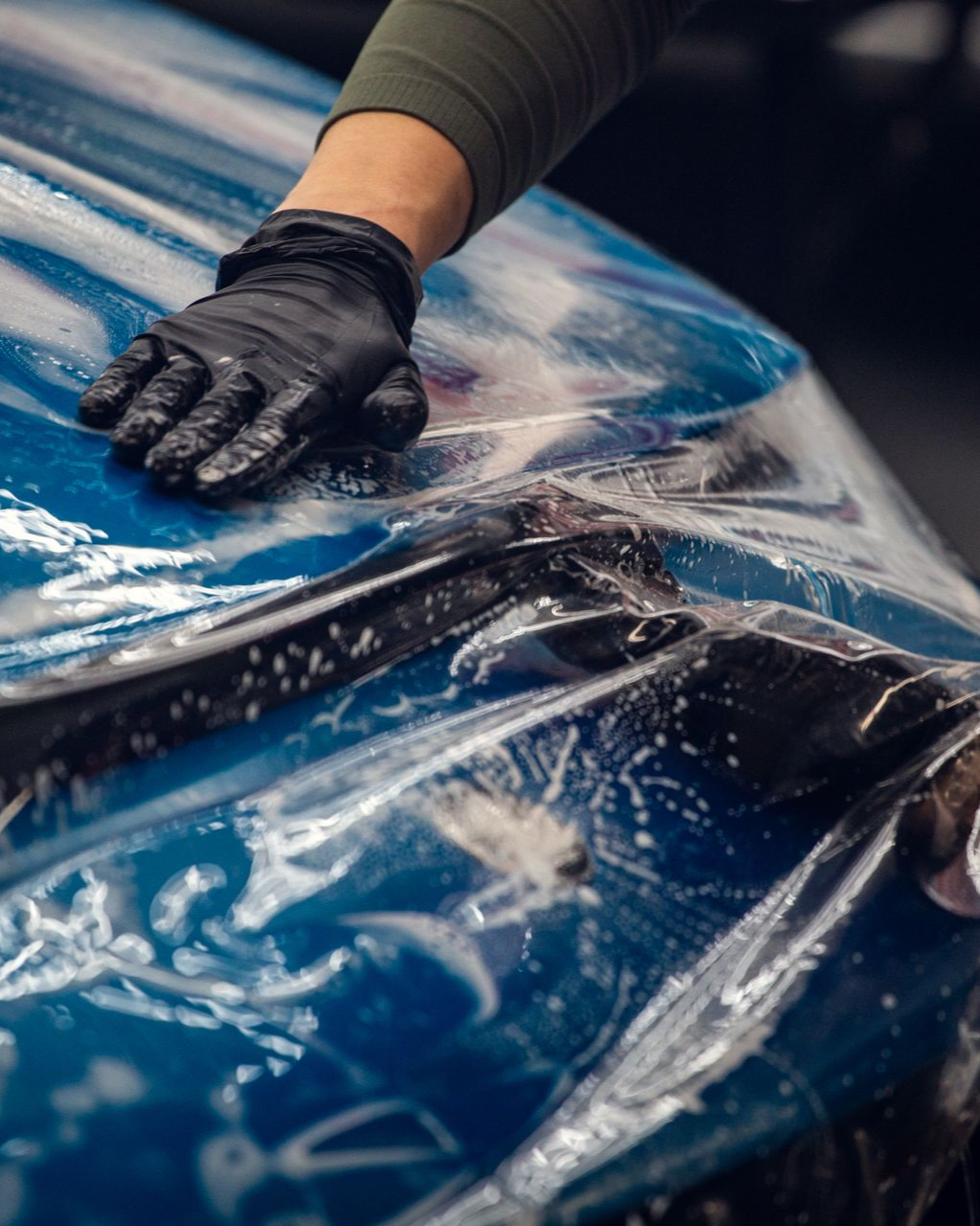 Gloved hand smoothing clear plastic film over a blue car hood in a workshop