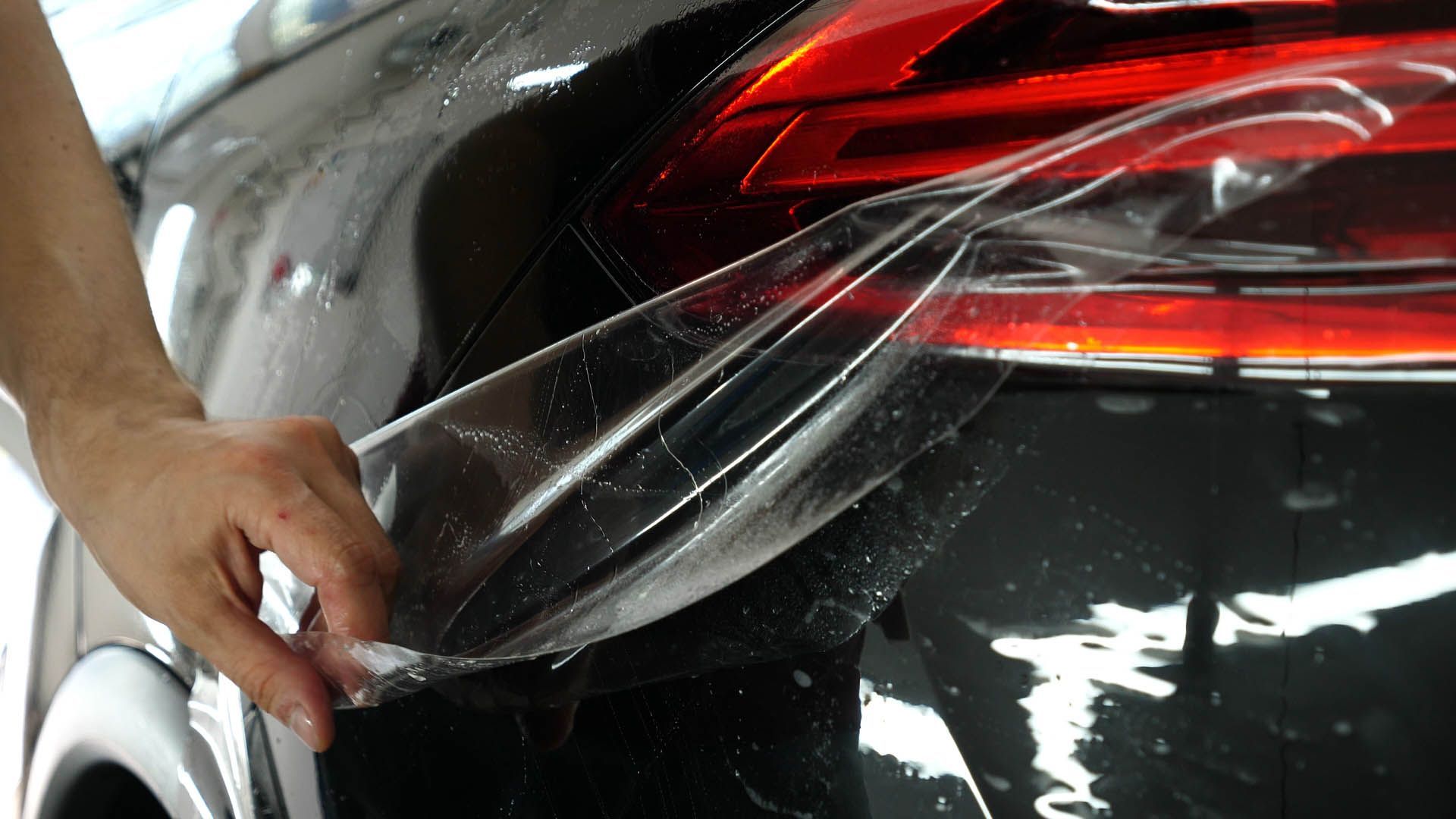 Hands applying clear protective film to a black car’s rear taillight area