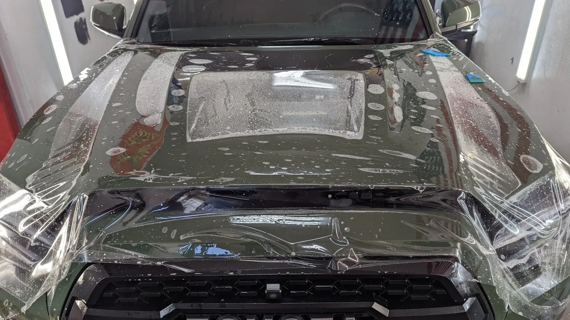 Car wrapped in clear protective film in a workshop, front view with hood and windshield covered.