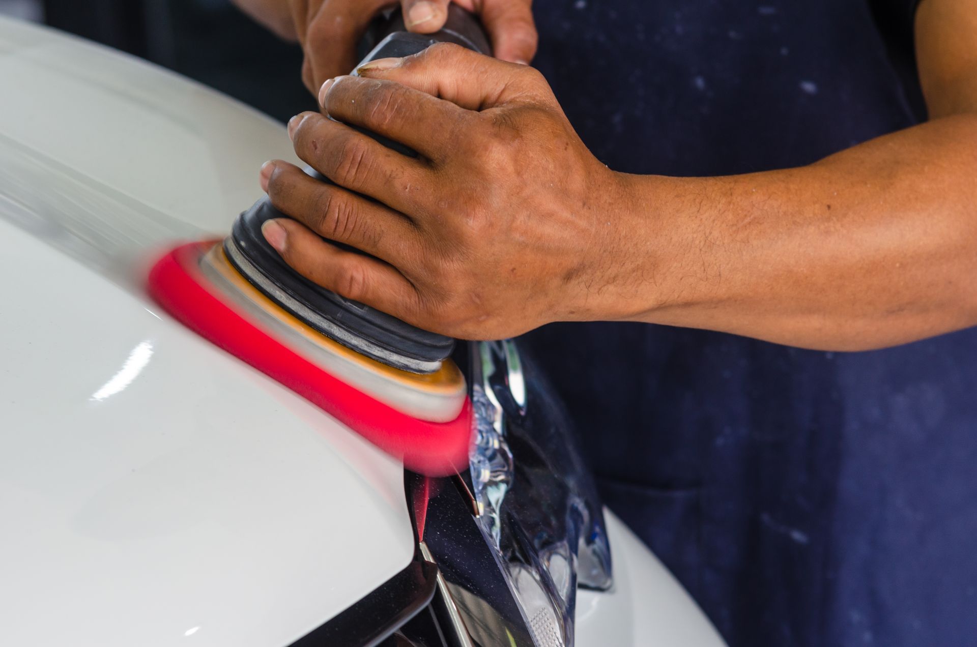 Hands polishing a white car with a buffer, creating red and gray swirl.
