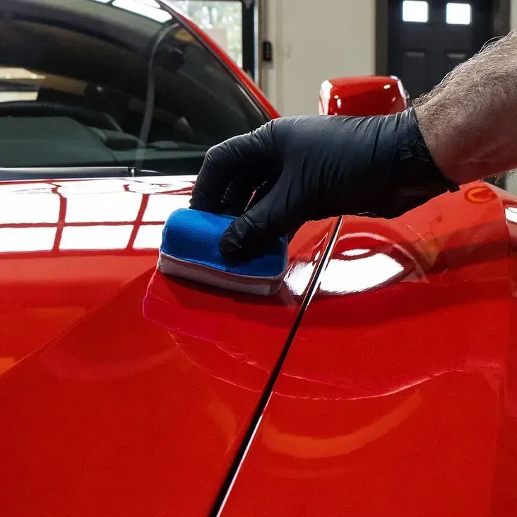 Gloved hand polishing a red car with a blue applicator sponge