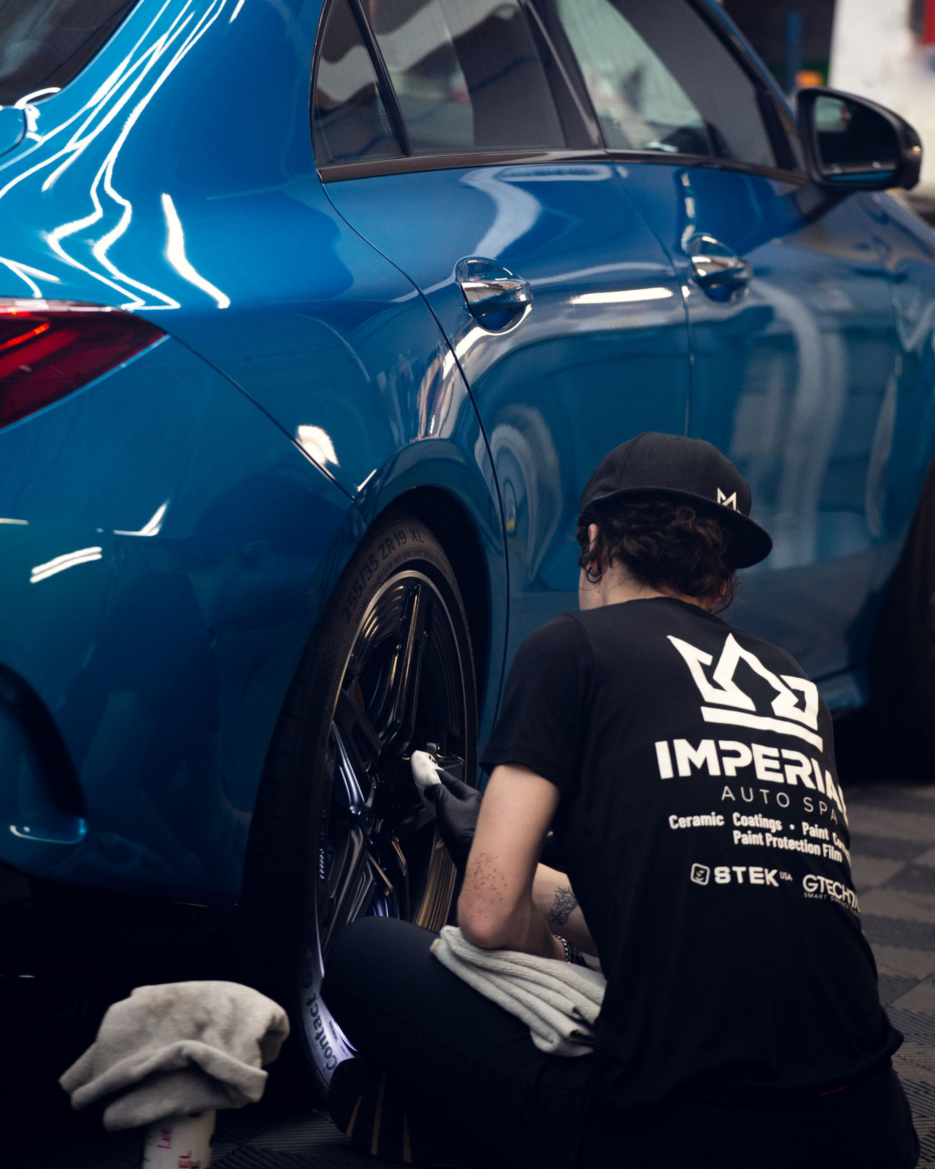 Person kneeling beside a blue car, cleaning the rear wheel area with cloths in a garage.