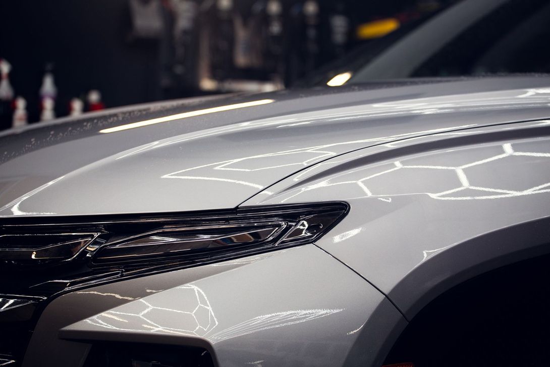 Close-up of a silver car’s front hood and headlight with reflections in a dim showroom