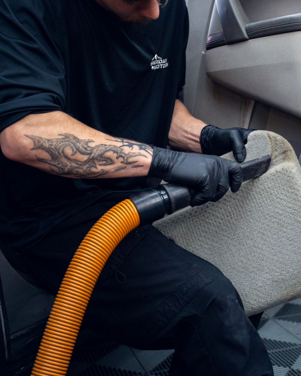 Person vacuuming gray car upholstery with a hose and brush inside a vehicle