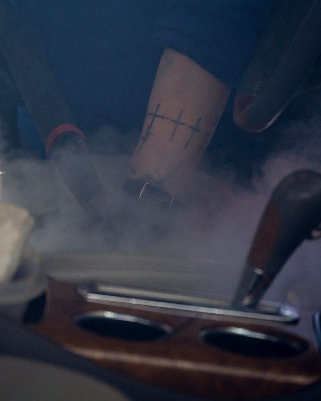 Close-up of a tattooed leg above a foggy engine with wooden trim and metal cup holders.