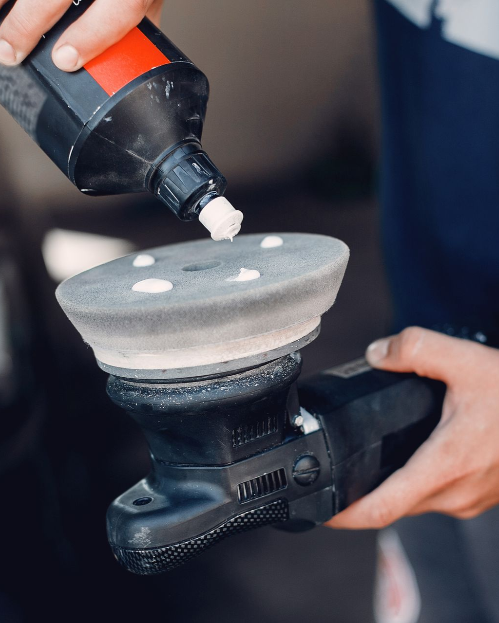 Hands apply polishing compound to a gray car buffer pad with a squeeze bottle.