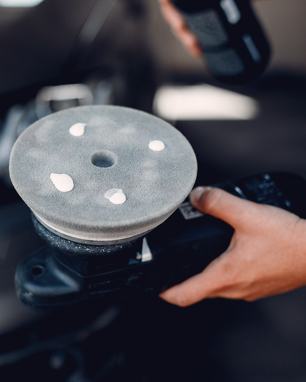 Hand holding a black orbital sander with a gray sanding pad and white spots.