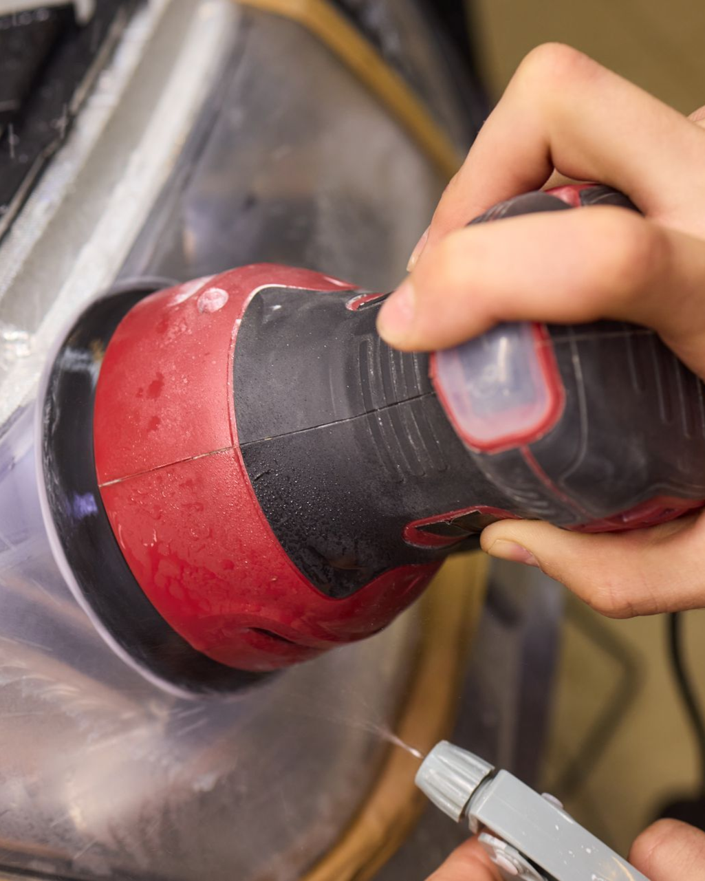 Hand sanding a red and black car part with a power sander, creating dust in a workshop