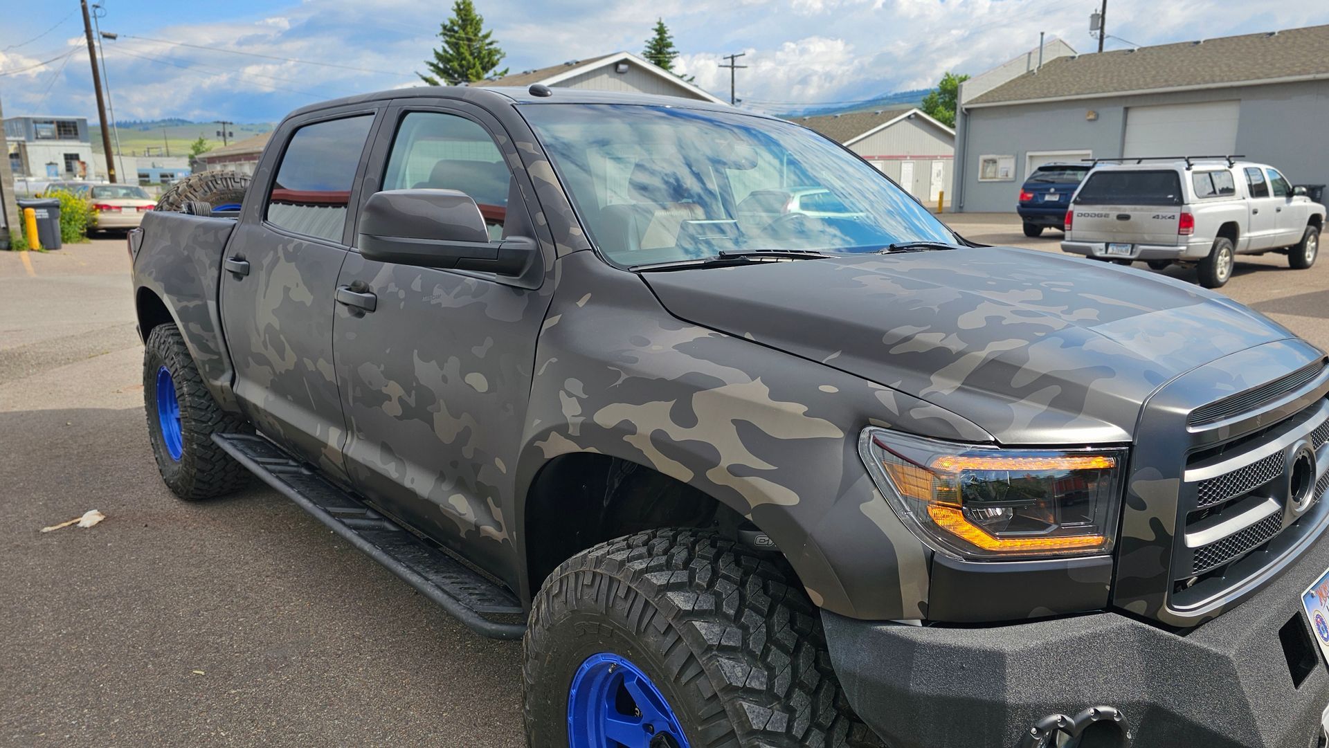 Camouflage-pattern pickup truck with blue wheels parked in a lot under a cloudy sky