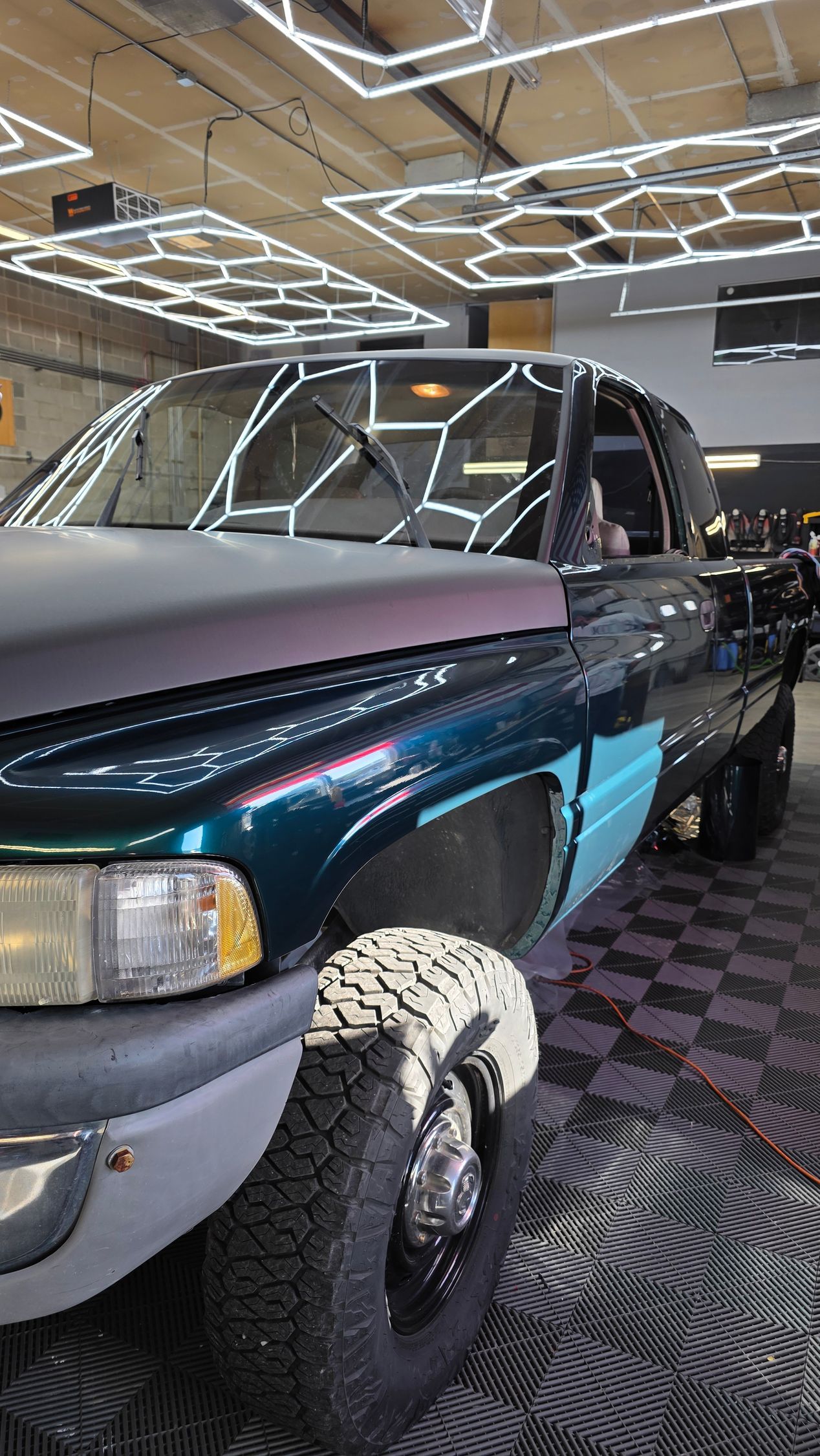 Dark teal pickup truck with a lifted front wheel displayed indoors under geometric ceiling lights.