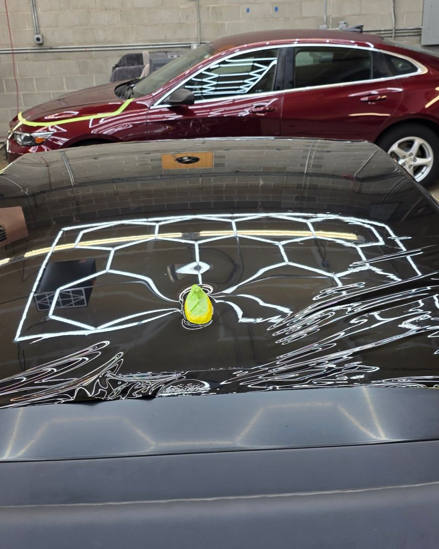 Black car hood reflecting a ceiling light pattern, with a yellow ornament near the windshield; maroon car in background