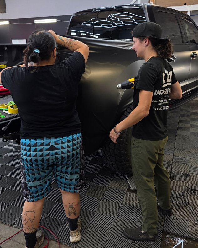 Two people detail a black SUV inside a workshop, with one polishing the hood and the other standing nearby.