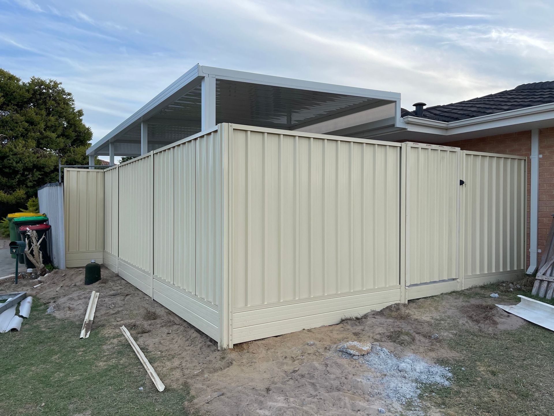 Beige corrugated metal enclosure beside a house, with a slanted roof and unfinished ground around it.
