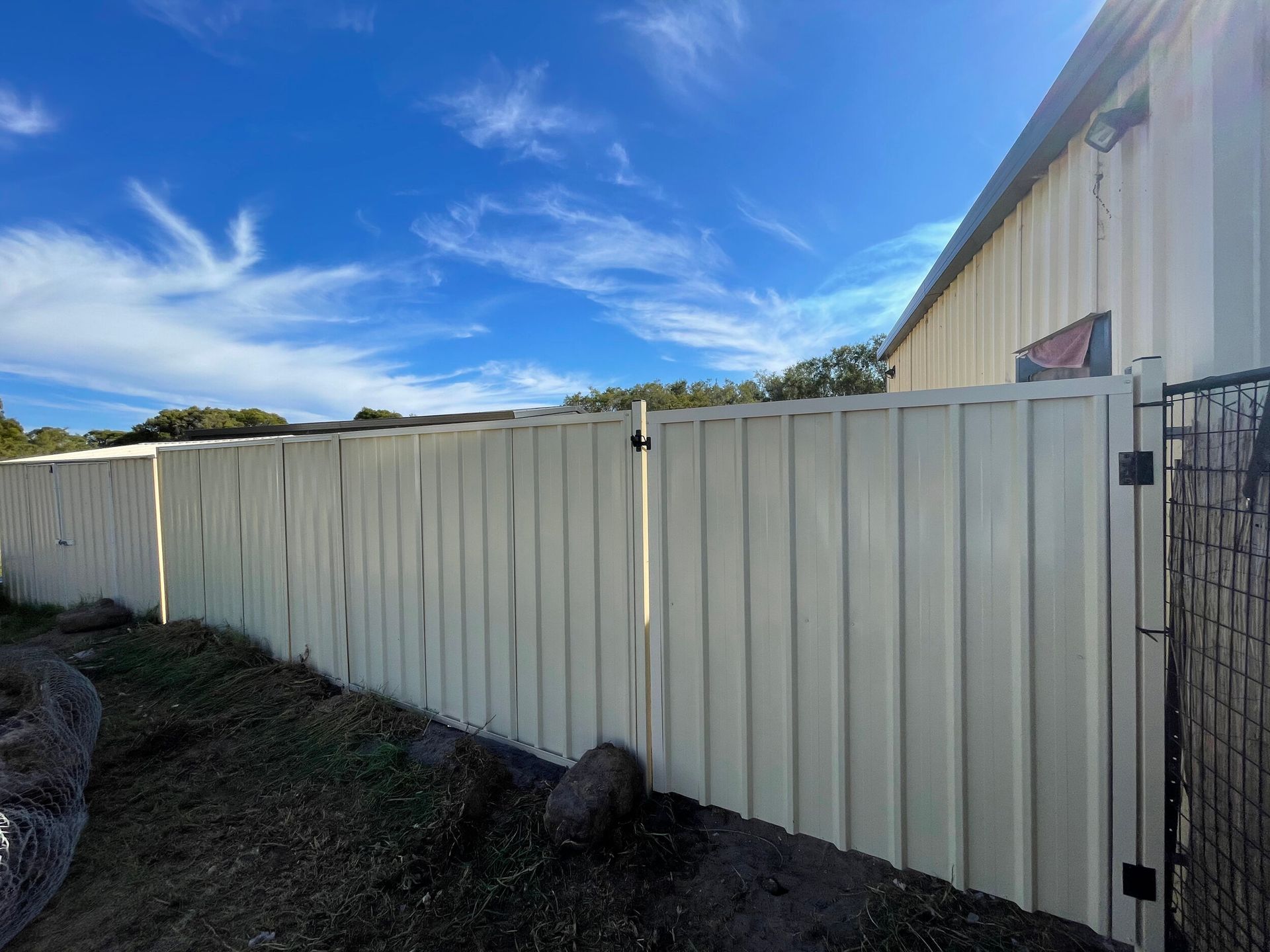 White corrugated metal fence beside a beige building under a blue sky with wispy clouds