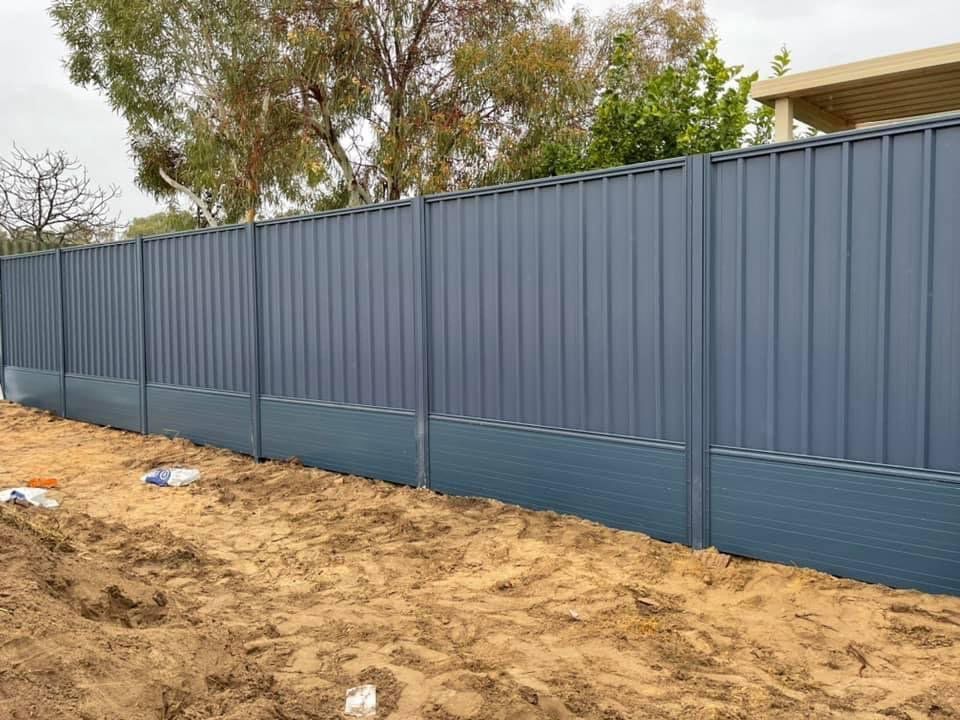 Blue corrugated metal fence along a dirt lot with trees in the background.