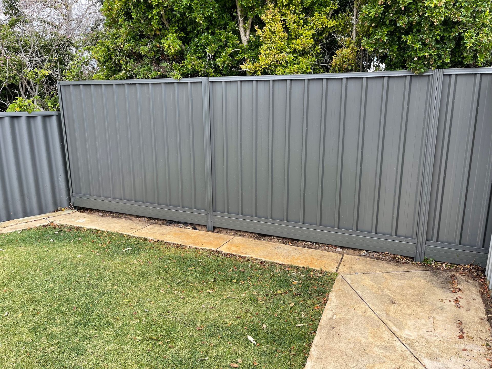 Gray metal fence beside a grassy yard and concrete path, with trees in the background.
