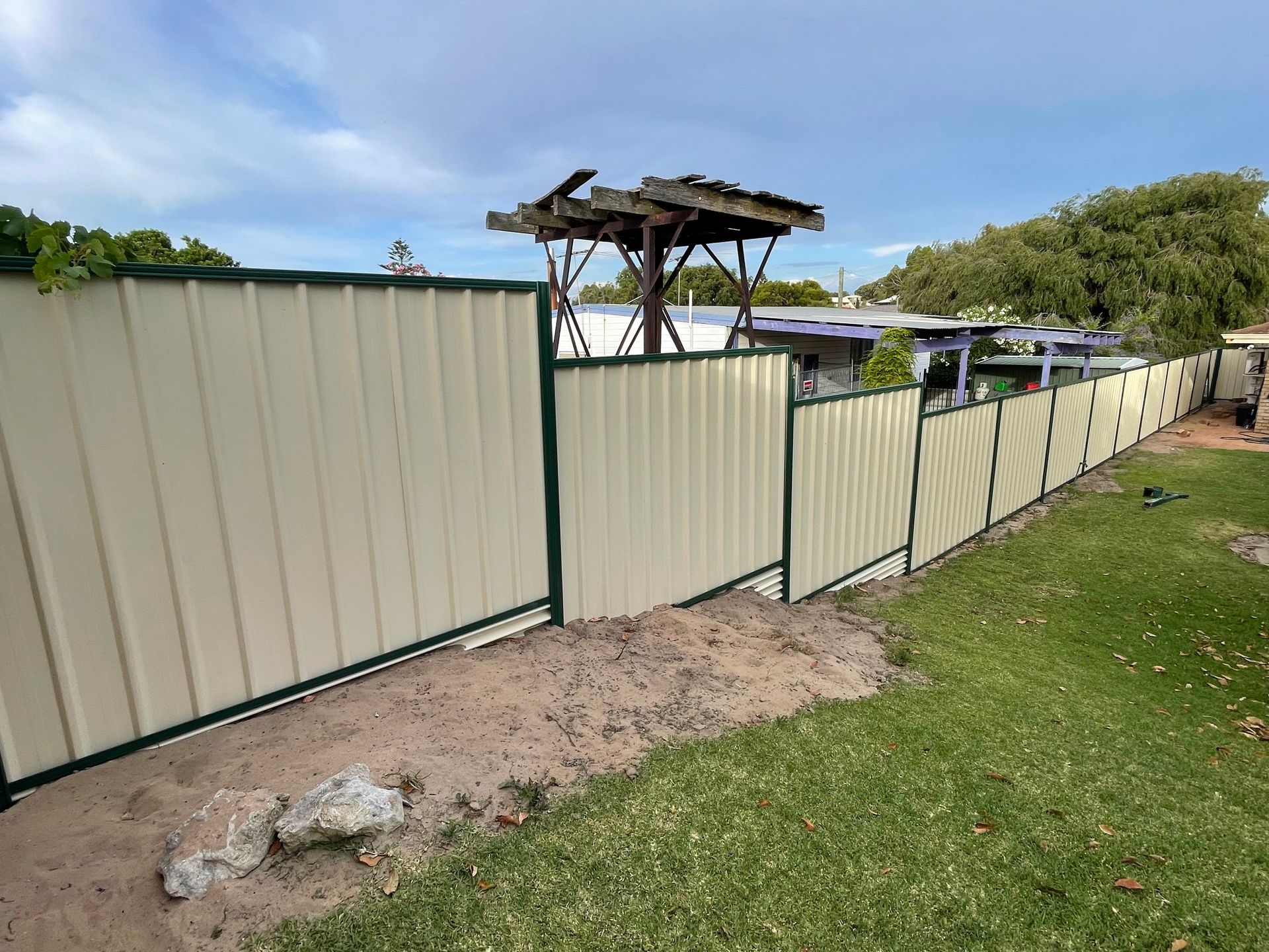 Corrugated metal fence along a grassy yard, with a small open wooden shelter above it.