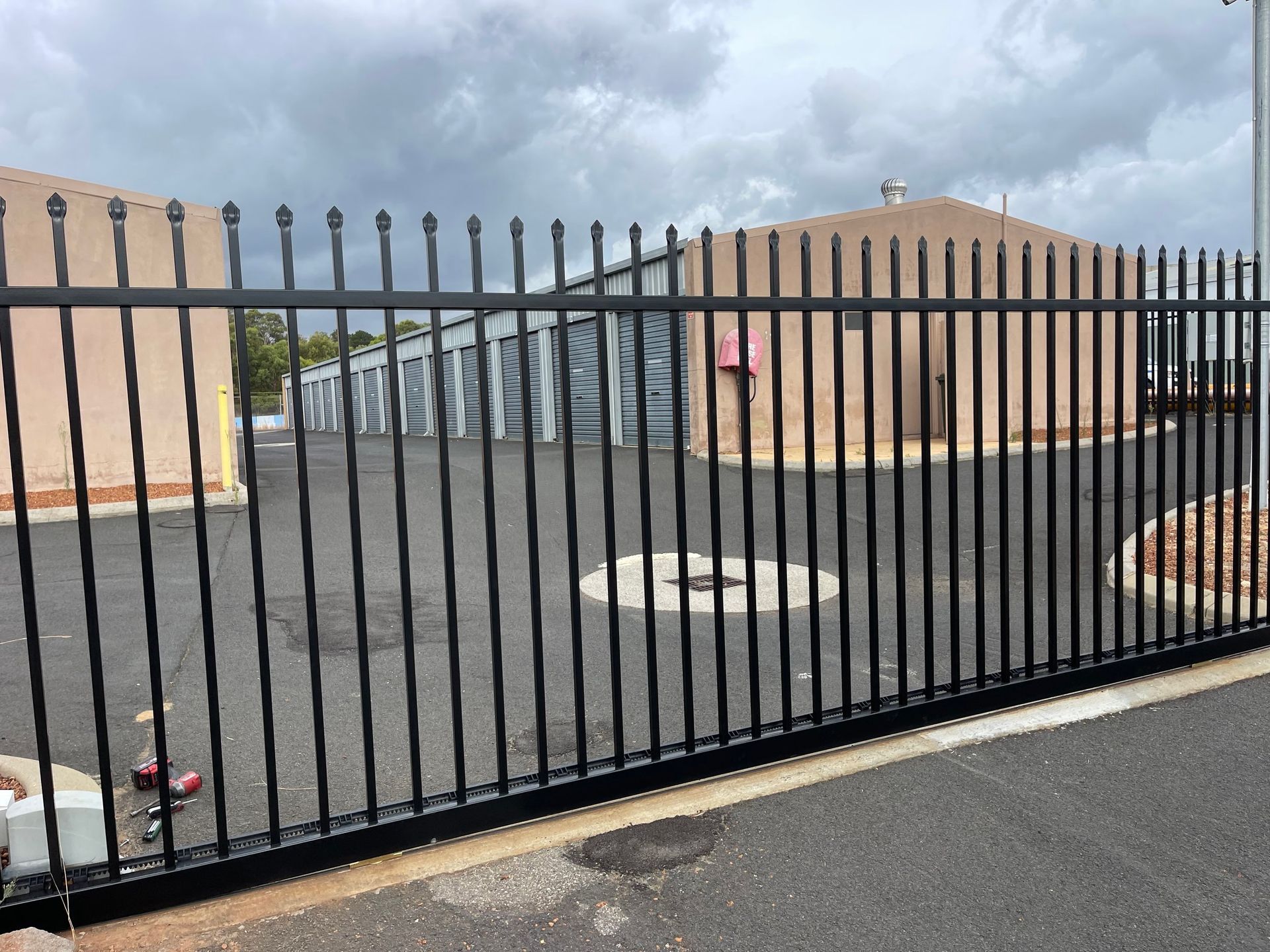 Black metal gate in front of a paved parking lot with beige buildings under a cloudy sky