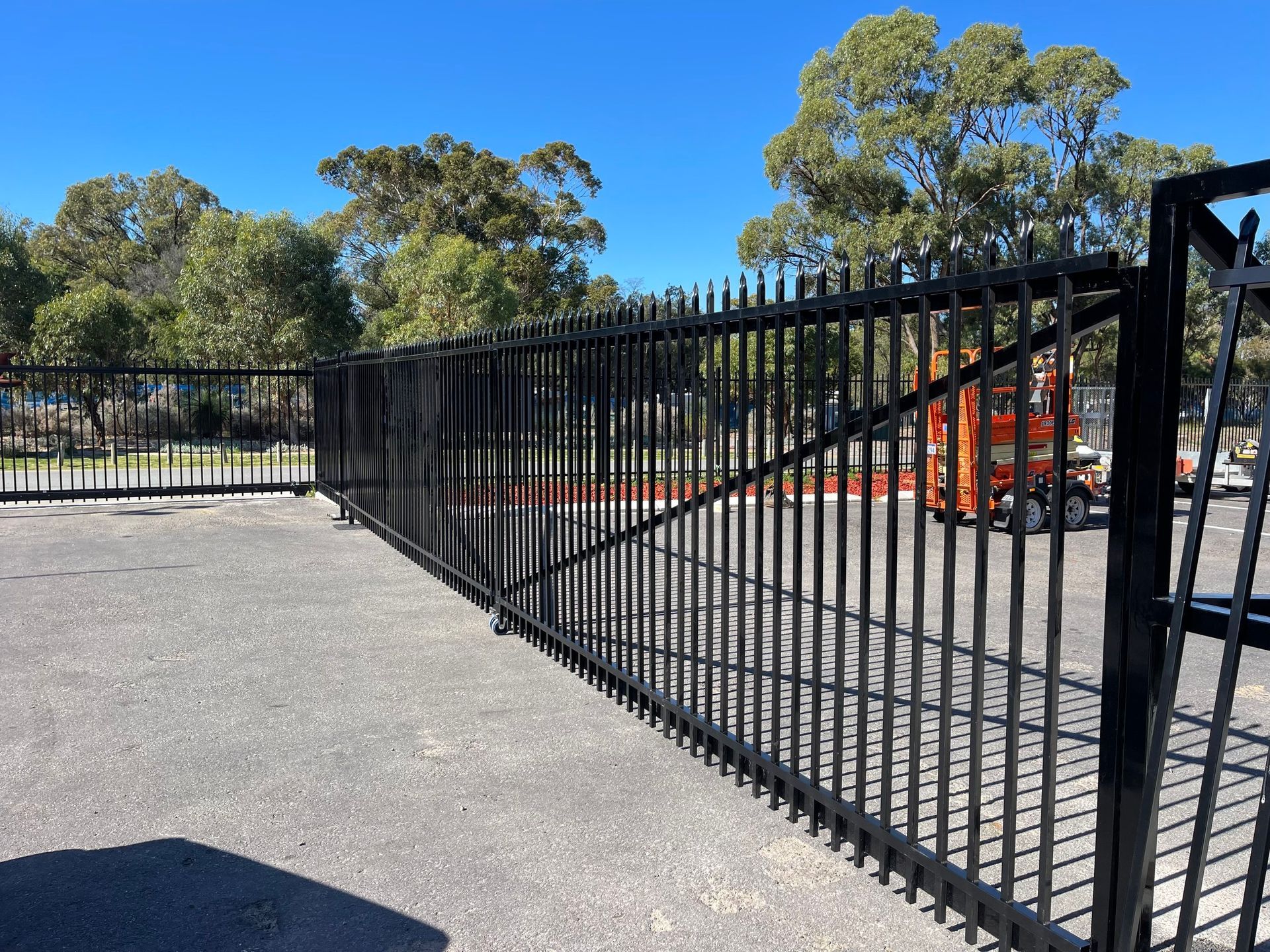 Black metal fence along a paved area with trees and orange construction equipment in the background