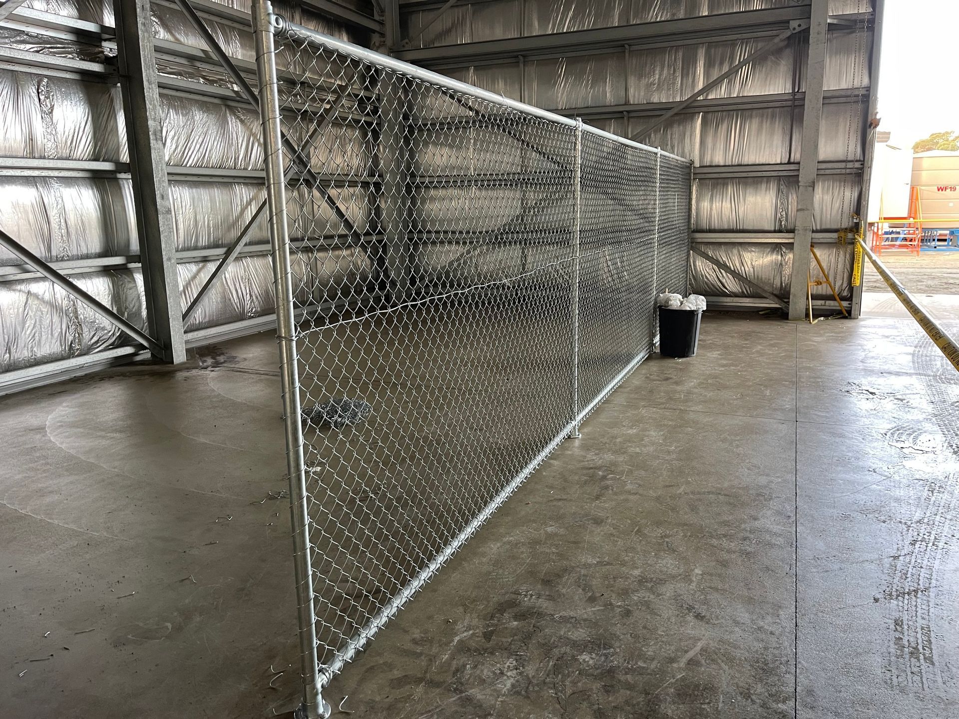 Chain-link fence panel in an industrial warehouse, with a black bucket on the wet concrete floor.