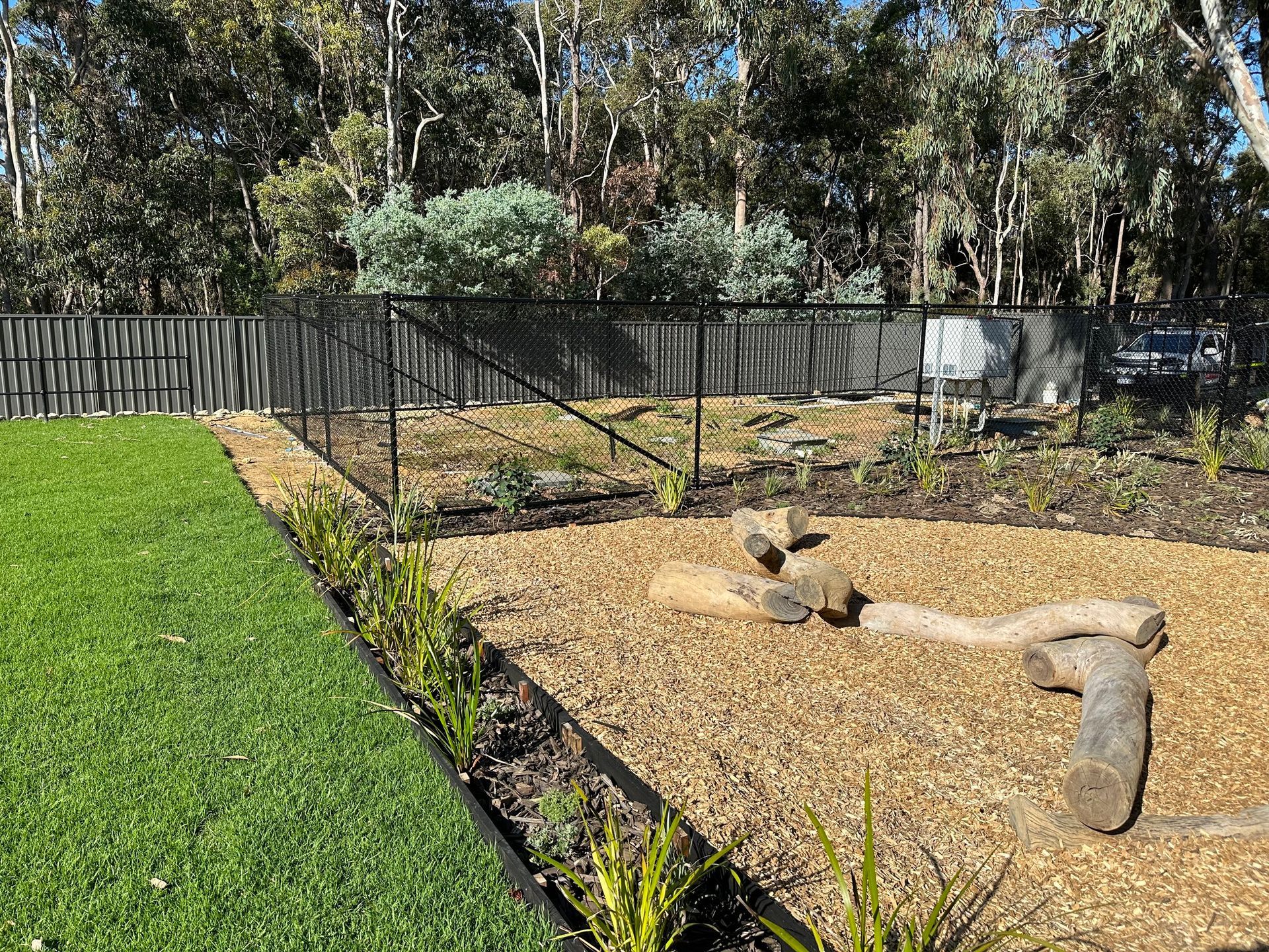 Backyard garden with mulch bed, trimmed grass, trees, and a black fence