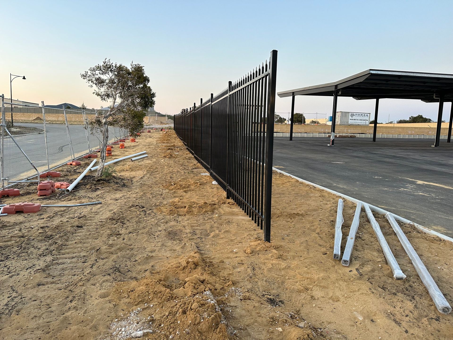 Chain-link fence along a dirt roadside beside a paved lot, with scattered pipes and open landscape at sunset
