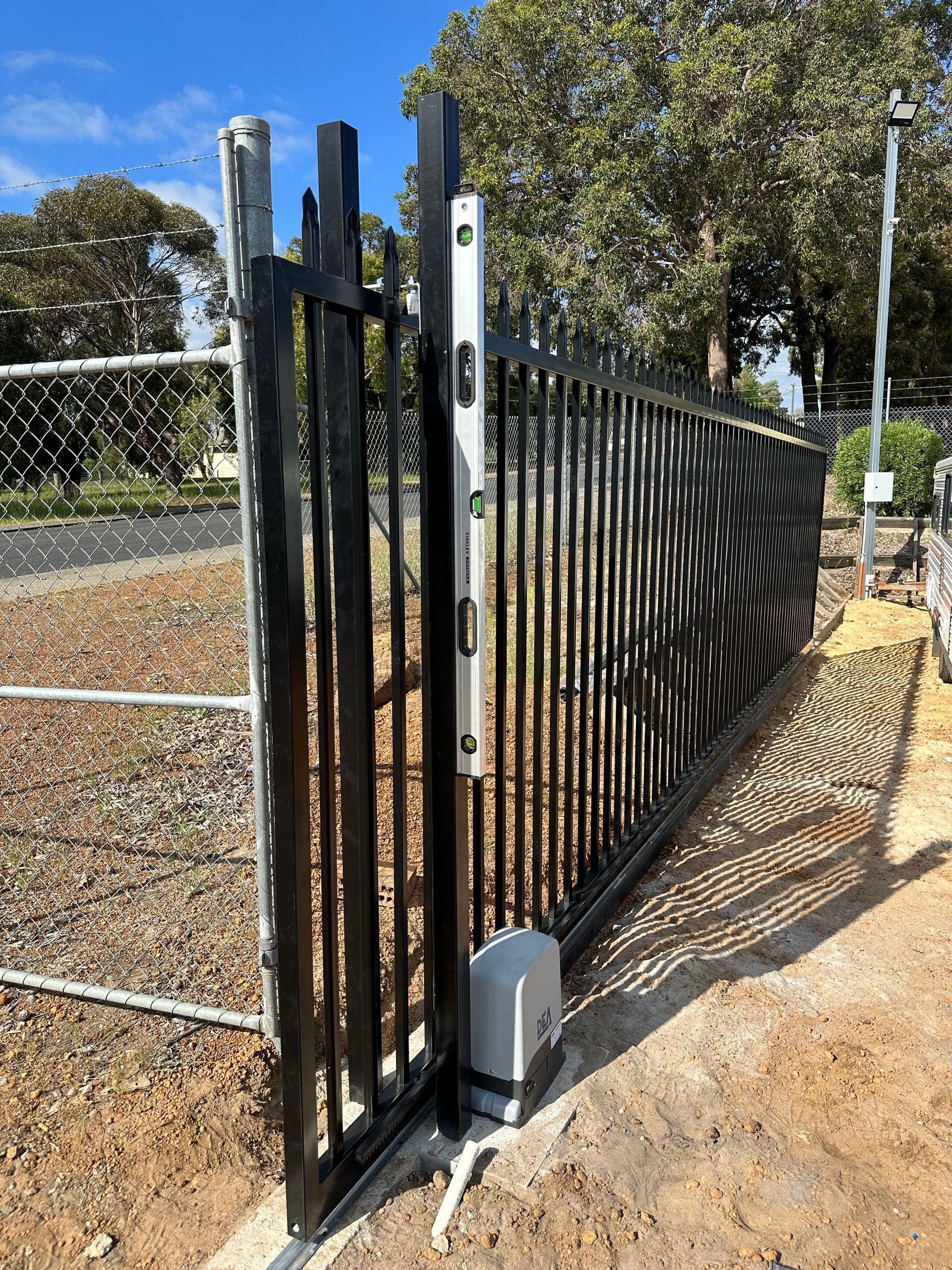Black metal sliding gate with a white post beside a chain-link fence in a sunny outdoor area