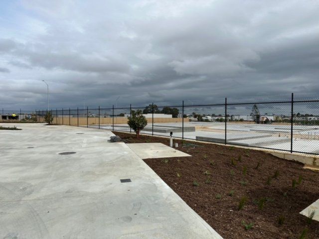 Empty concrete lot with a chain-link fence and cloudy sky beside a landscaped strip.