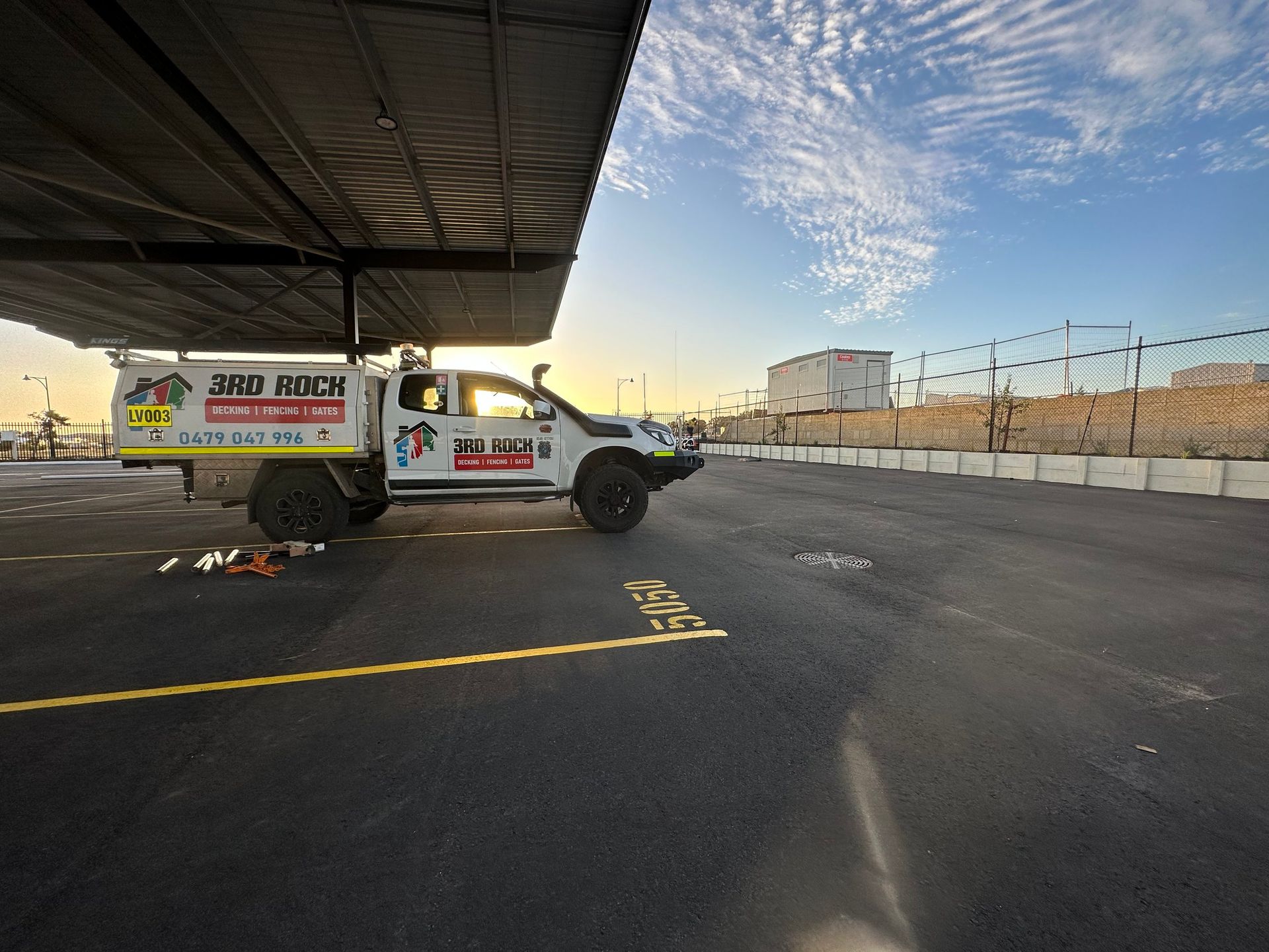 White pickup with advertising trailer parked in a lot under a canopy at sunset, beside a chain-link fence.