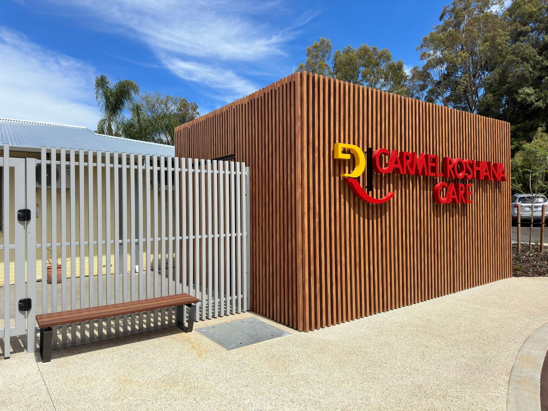 Modern building with vertical wood slats, red sign, white gate, and bench under a blue sky