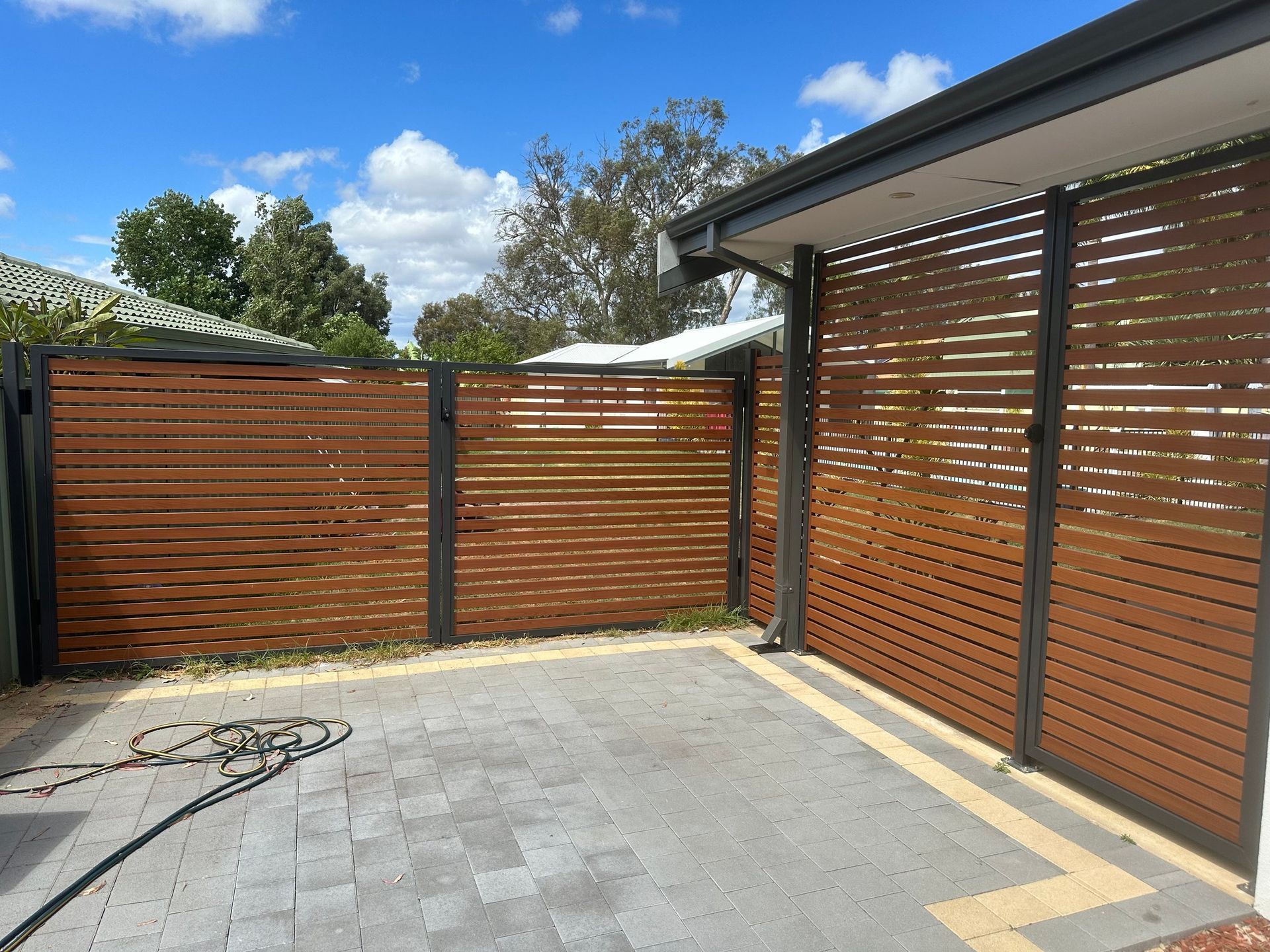 Paved backyard patio with wood privacy fencing, black gate, and house wall under a blue sky