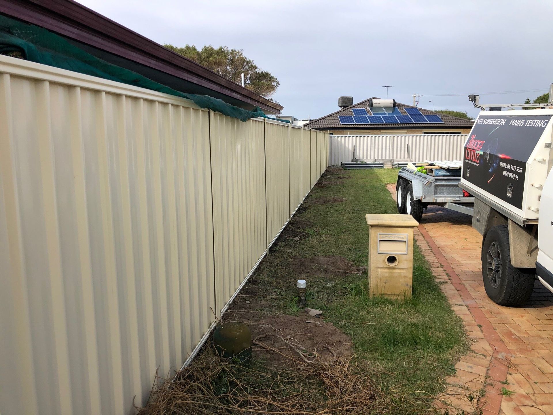 Narrow side yard with cream fence, grass strip, brick path, mailbox, and parked white utility vehicle