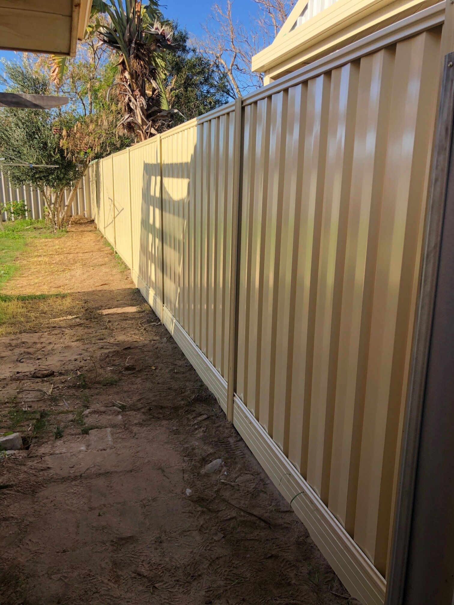 Beige corrugated metal fence along a narrow dirt path beside a house in daylight