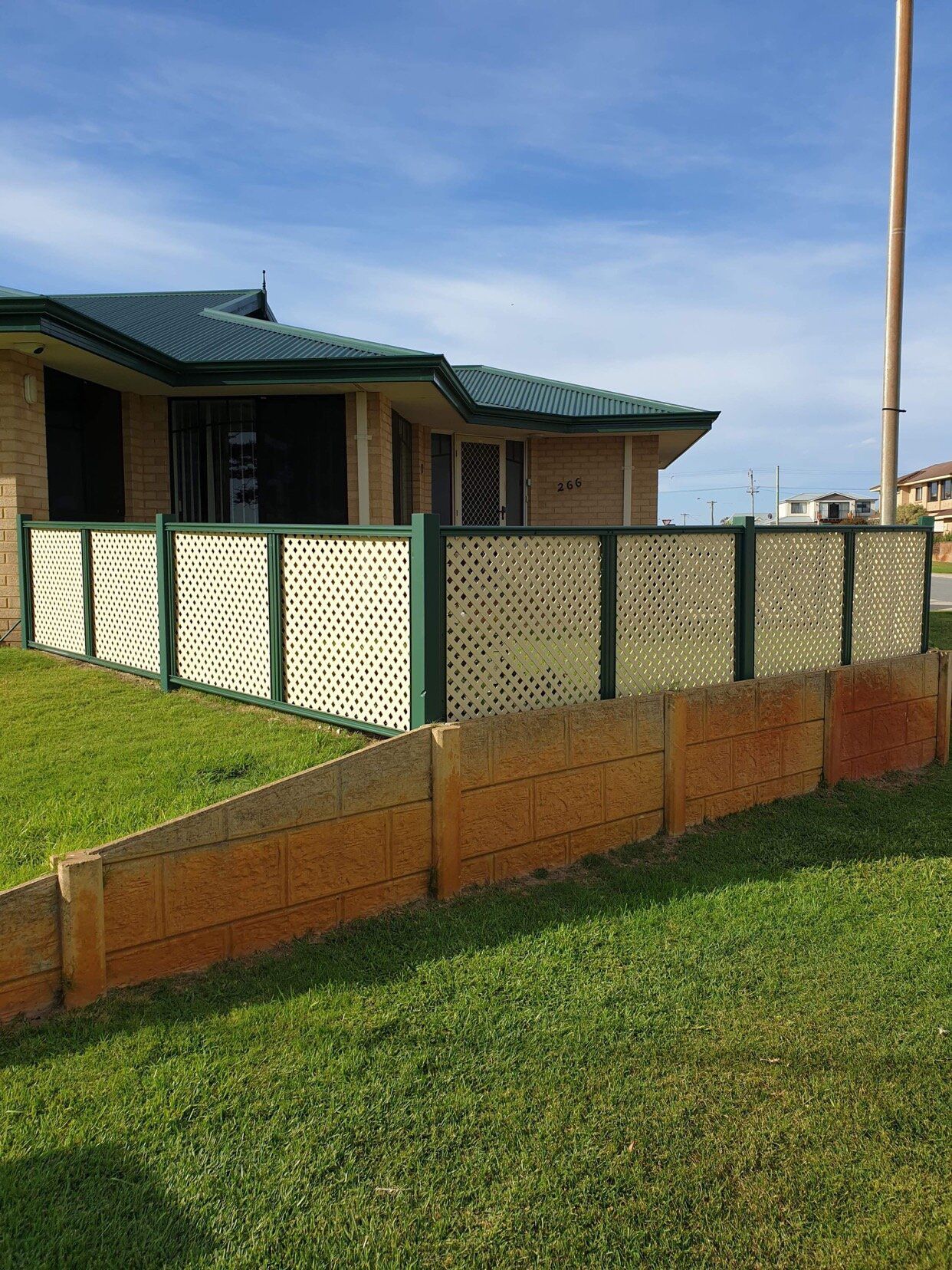 Suburban house with green roof, fenced yard, and a sloped brick retaining wall beside a grassy lawn
