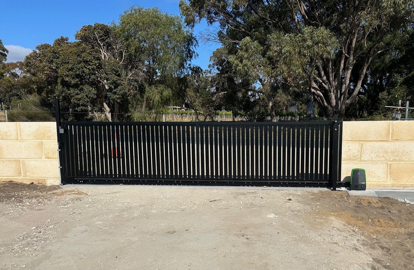 Black metal sliding gate between beige stone walls with trees behind and a gravel driveway in front