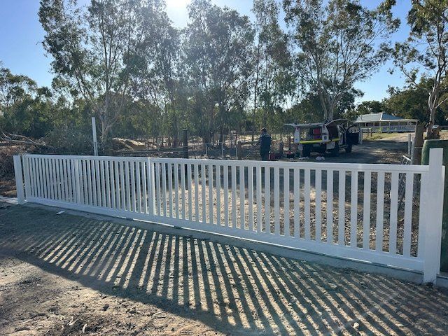 White metal fence beside a sunny dirt road, with trees and parked vehicles in the background.