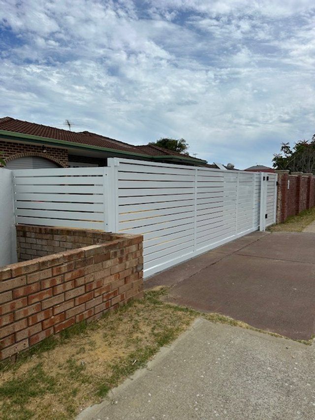 White slatted fence beside a brick wall and driveway under a cloudy sky